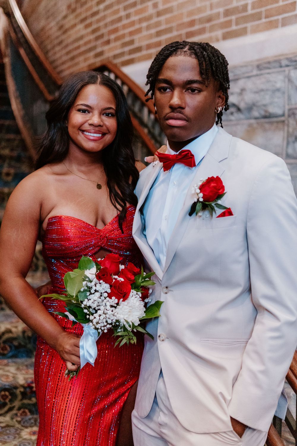 A couple poses together at a formal event, with one wearing a sparkling red dress and the other in a white suit.
