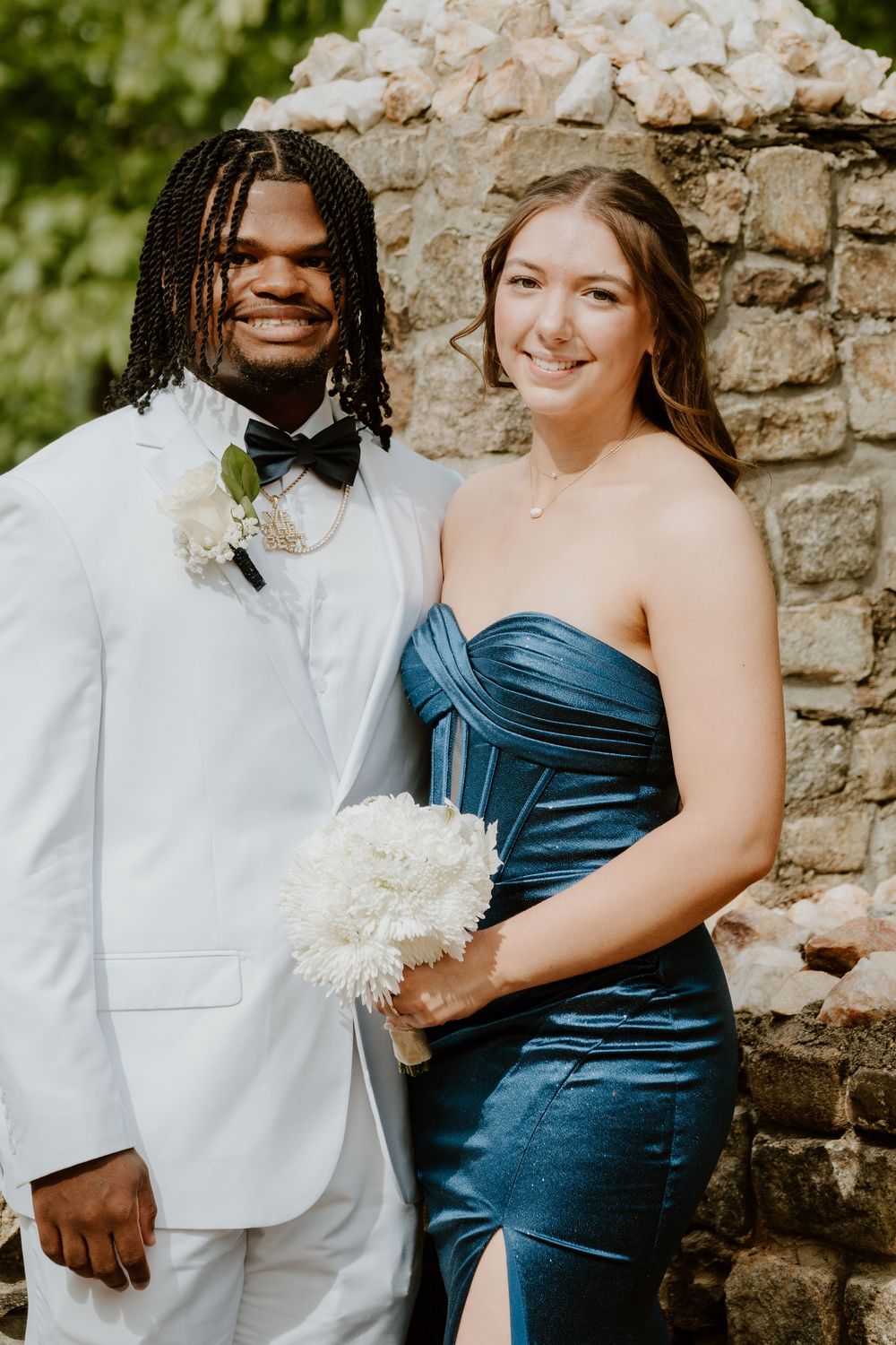 Couple in formal wear poses against stone wall backdrop at outdoor event.