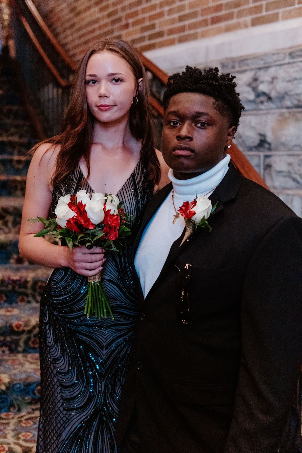 A formal couple poses together against a brick wall backdrop, one in black and one in formal attire.