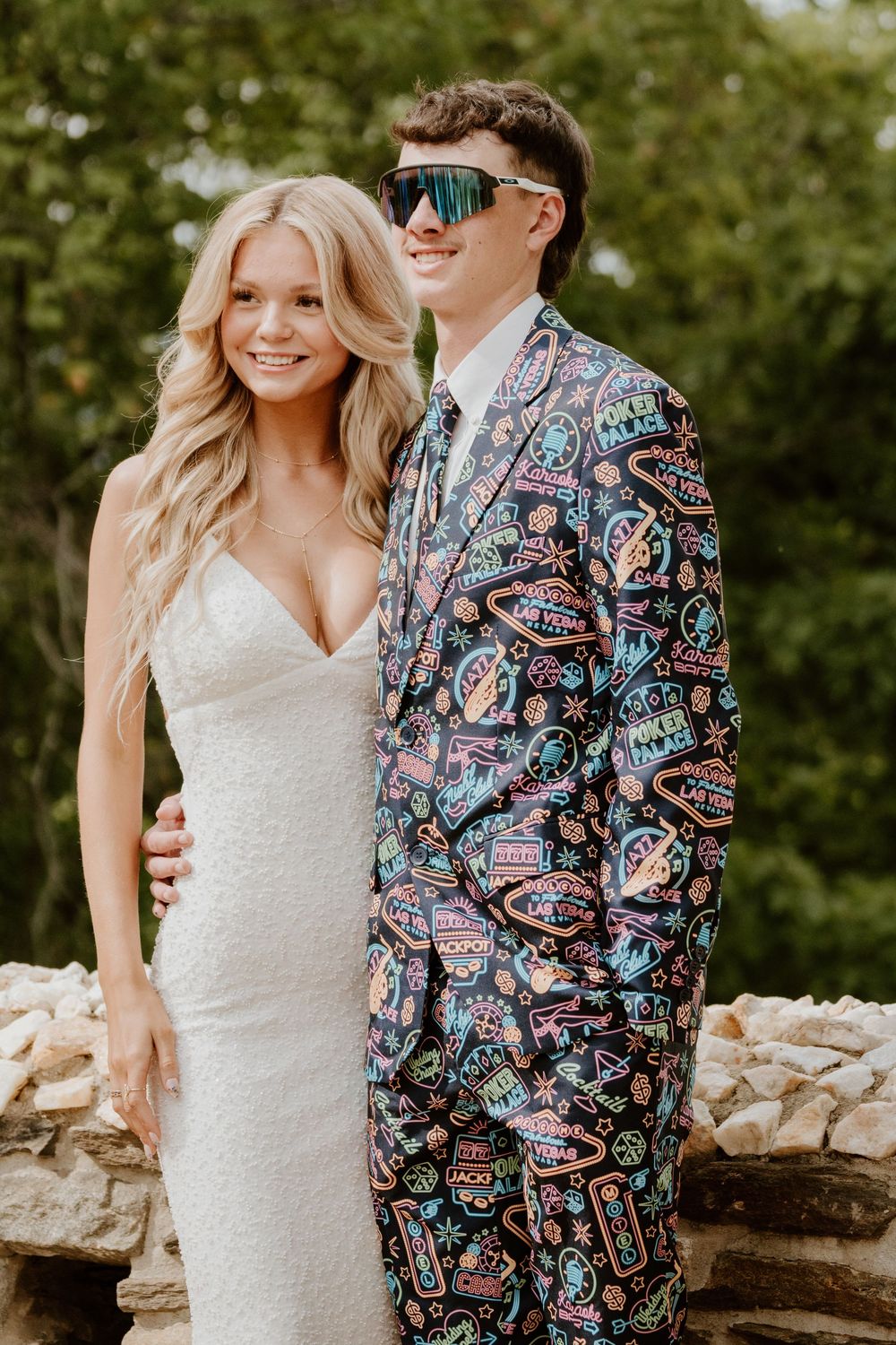 Couple poses outdoors, with white dress and colorful patterned suit jacket.