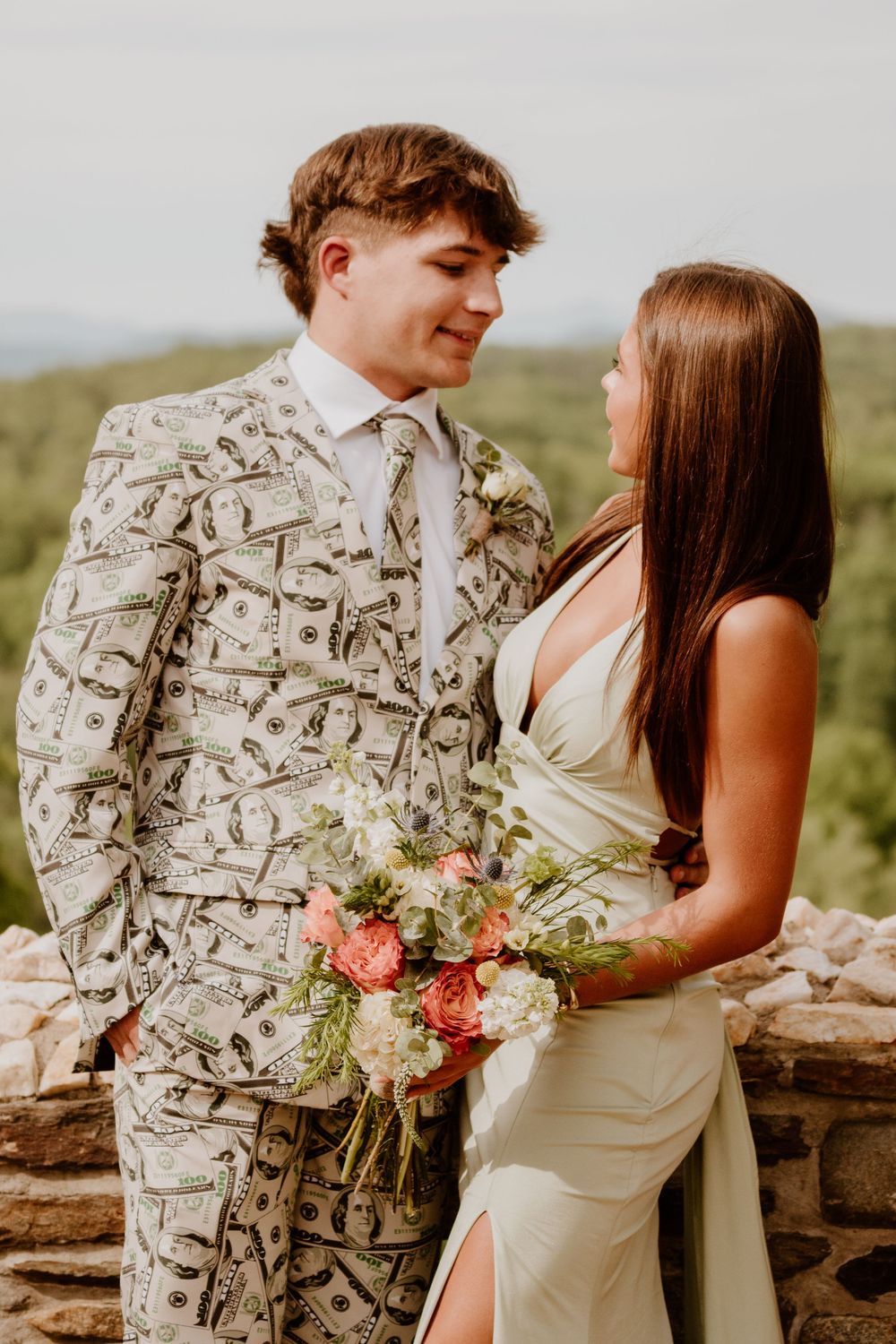 Couple in formal wear poses by stone wall with floral bouquet.