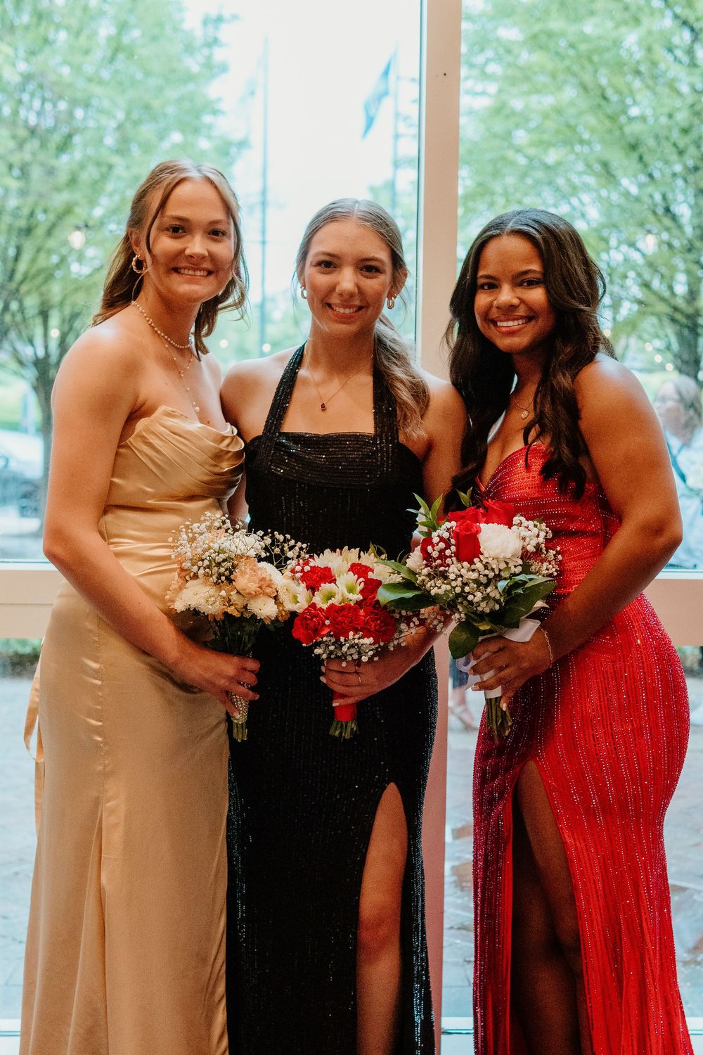 Three friends in formal dresses holding bouquets at a special event.