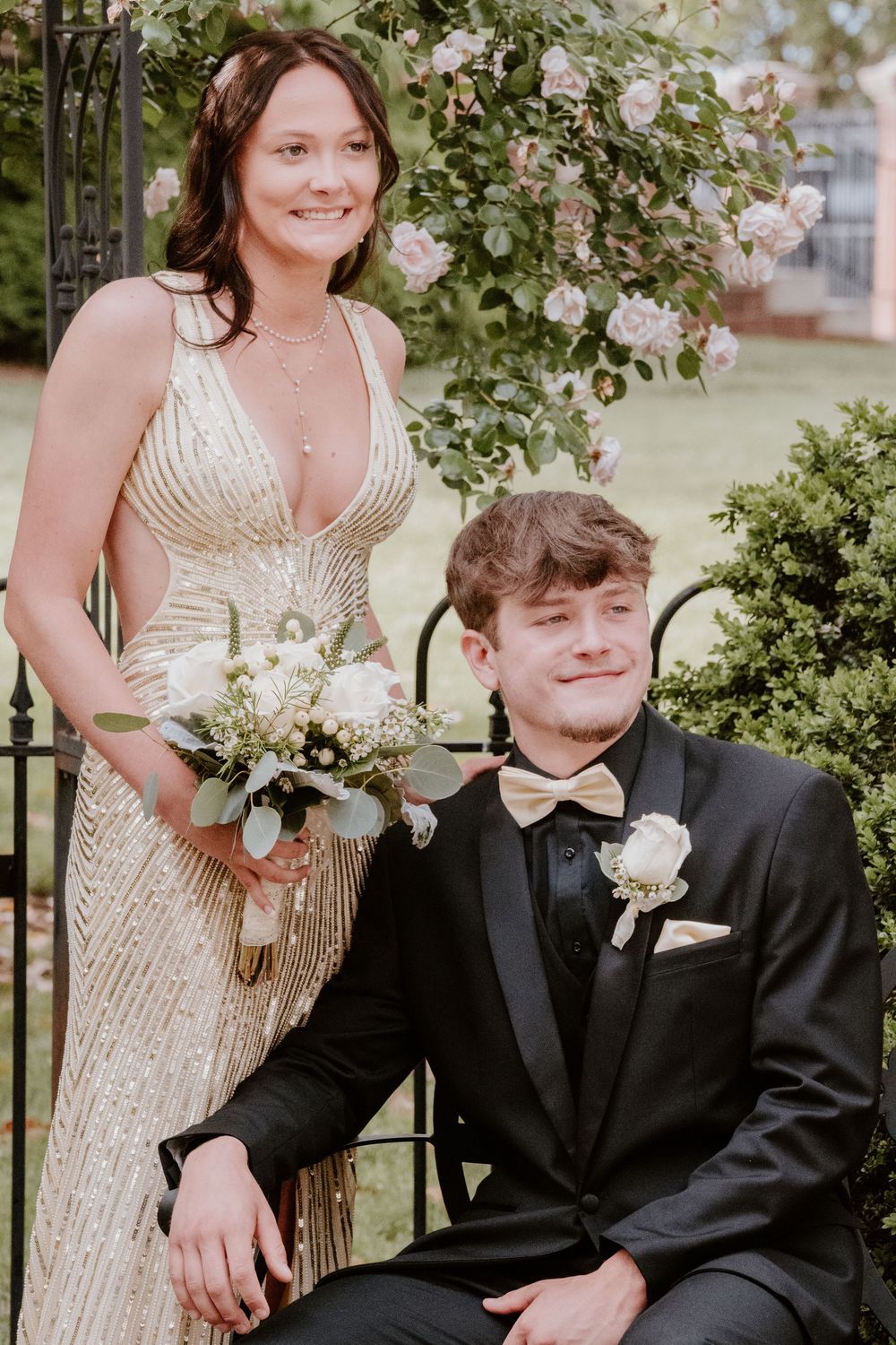 Couple in formal attire poses together under tree branches with gold sequined dress and black suit.
