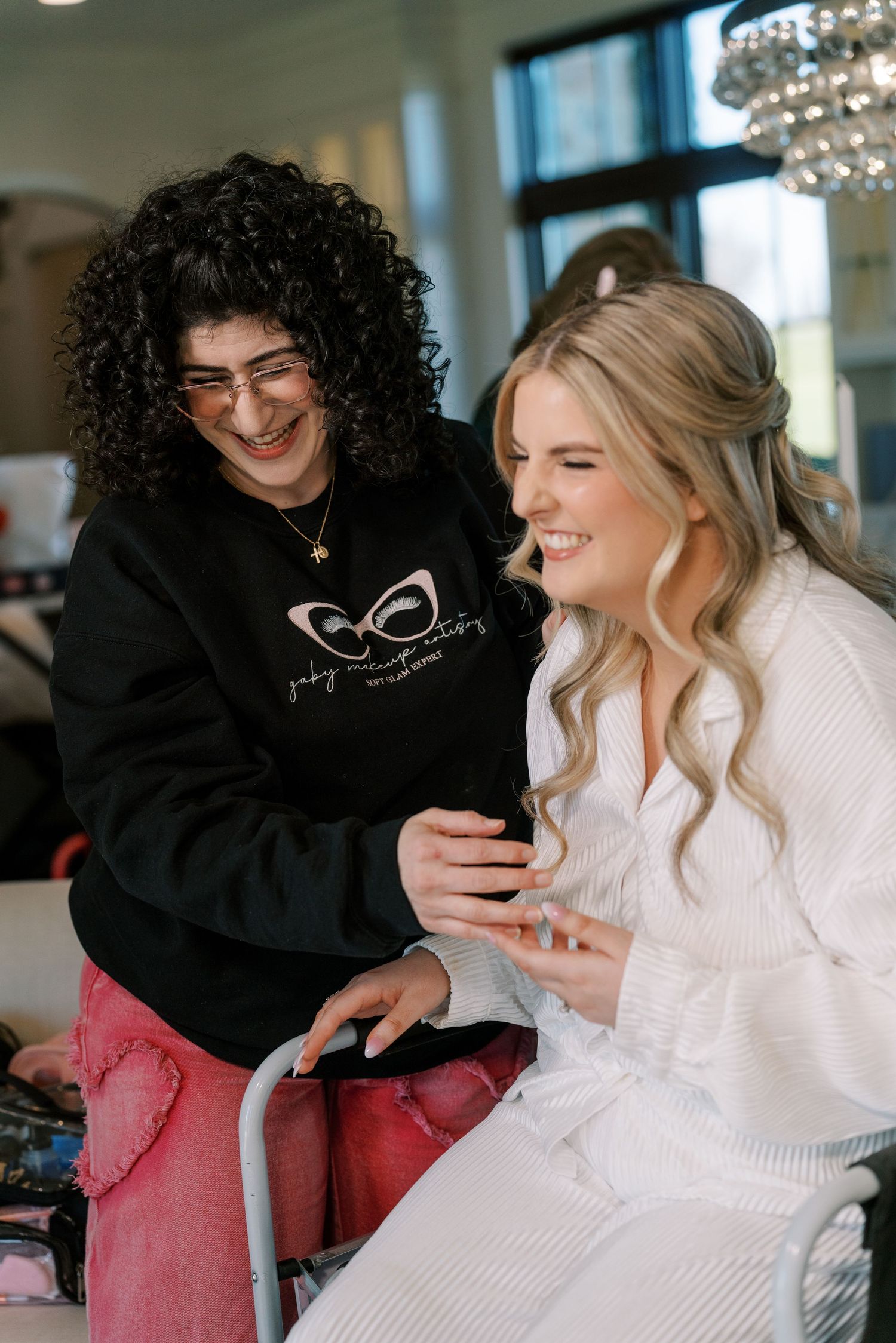 A makeup artist in okc applies makeup to a bride on her wedding day. They are smiling and have fun/