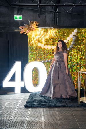 Woman in elegant gray gown poses beside illuminated number 40 at a glamorous birthday celebration with gold sequin backdrop.
