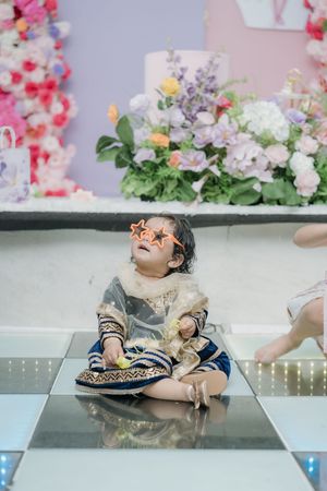 A small child in festive attire sits on a reflective surface with colorful flowers in the background.