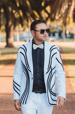 A person wearing a stylish white blazer with black trim and sunglasses poses outdoors in warm sunlight.