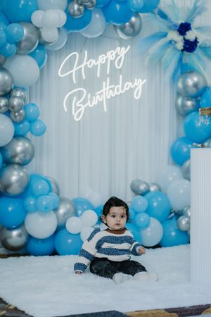 Baby boy sitting in front of a Happy Birthday neon sign with blue, silver, and white balloon arch backdrop.