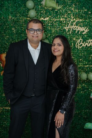 A couple in formal attire smiling at a birthday celebration with a green floral backdrop and neon Happy Birthday sign.