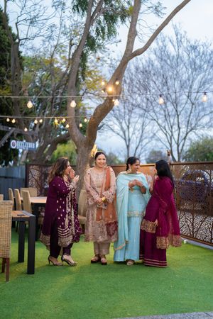 Four women in colorful traditional South Asian attire gather outdoors at a festive evening event with string lights.