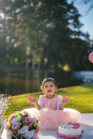 A joyful cake smash photo session outdoors with a pink tutu dress and floral decorations on a grassy hill.