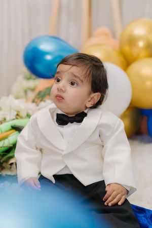 Small child wearing formal white tuxedo jacket and bowtie sits among colorful balloons at a celebration.