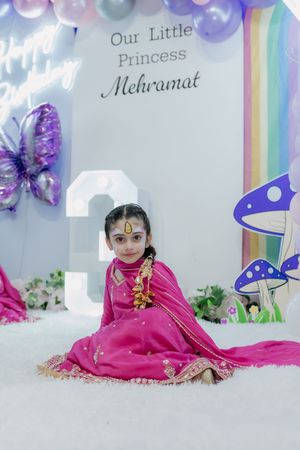 Young girl in pink traditional dress sits at her 3rd birthday party with princess Mehramat balloon backdrop.