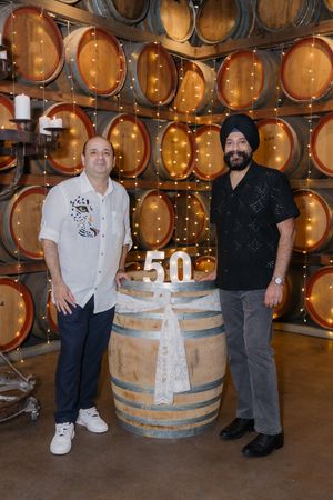 Two people pose in front of wooden wine barrels in a cellar or winery storage area.
