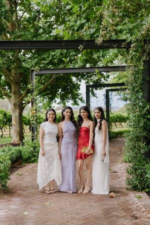 Wedding party in pastel dresses poses beneath lush green trees on a garden path.