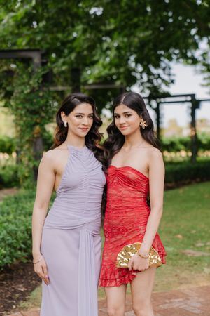 Two women in contrasting lavender and red dresses pose together in a garden setting for formal photos.