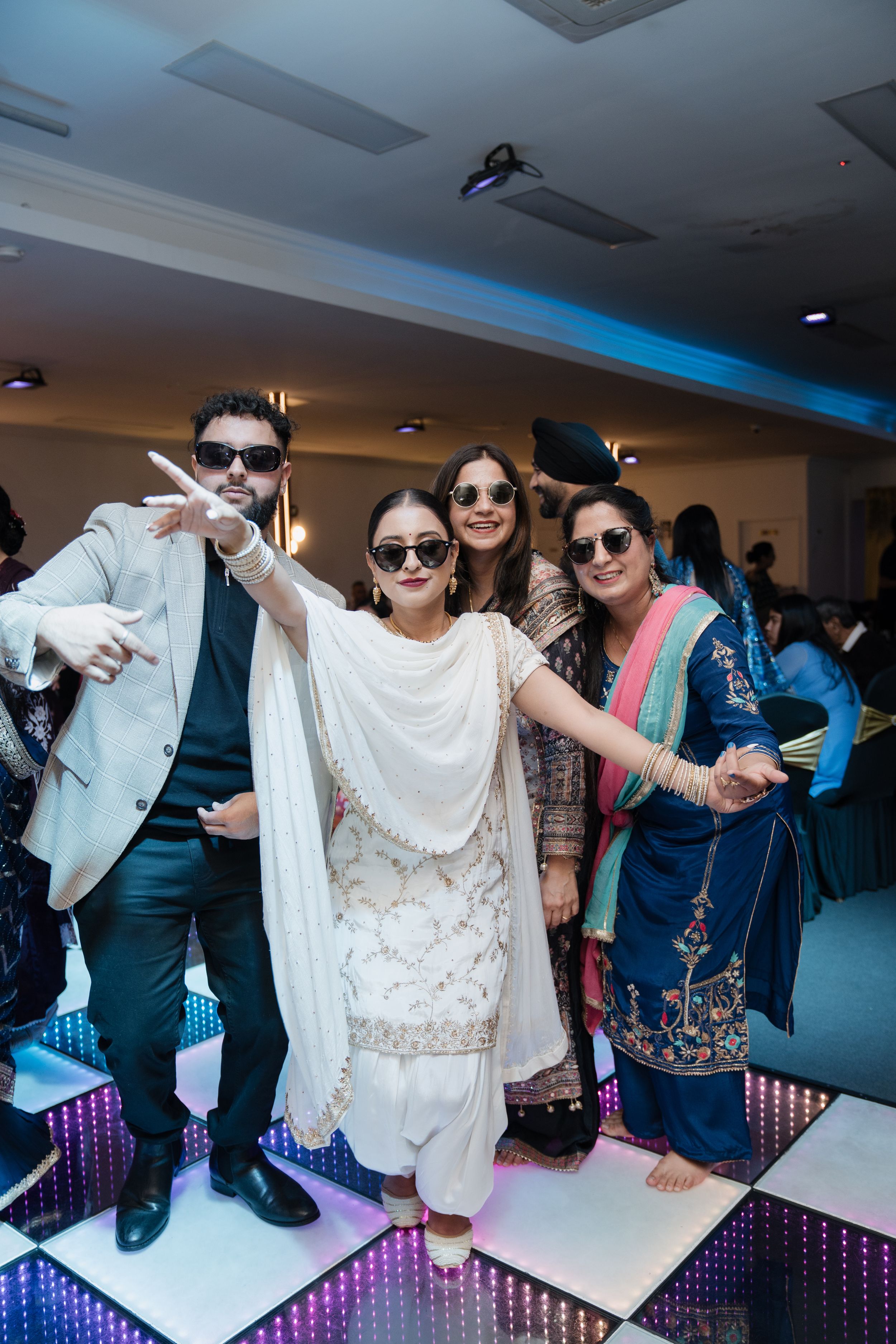 Group of friends in stylish traditional Indian attire pose and smile on an illuminated dance floor at a celebration.