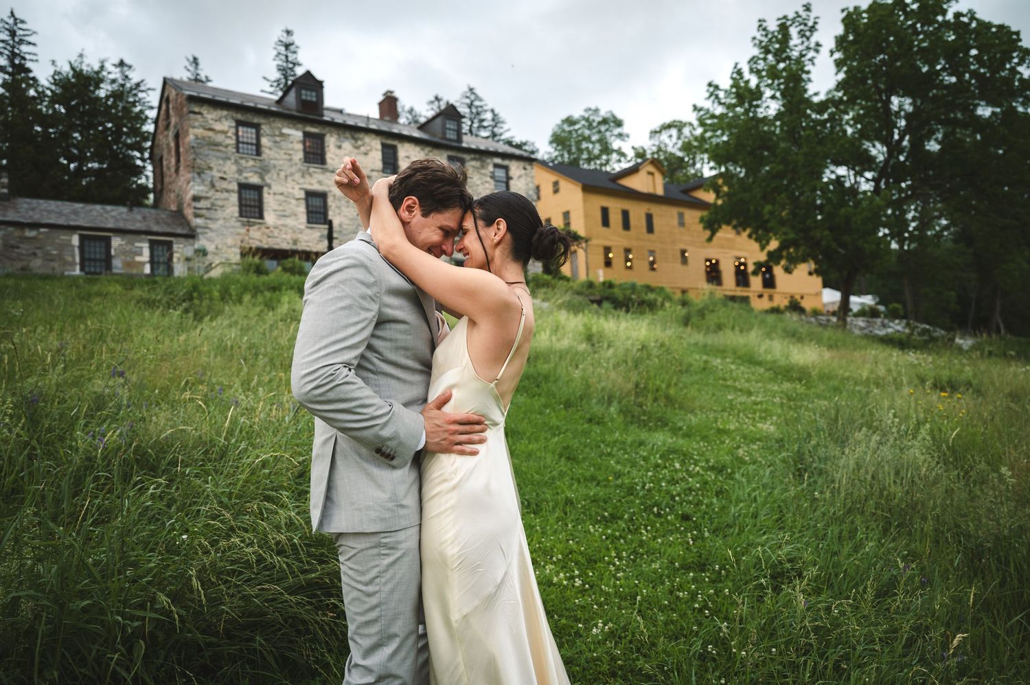 Wedding couple's romantic embrace on a grassy hillside with rustic stone buildings at The Ruins at Sassafras in New Lebanon NY Hudson Valley
