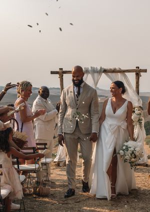 Outdoor celebration event with people in white and beige attire gathered under wooden beams against a natural backdrop.