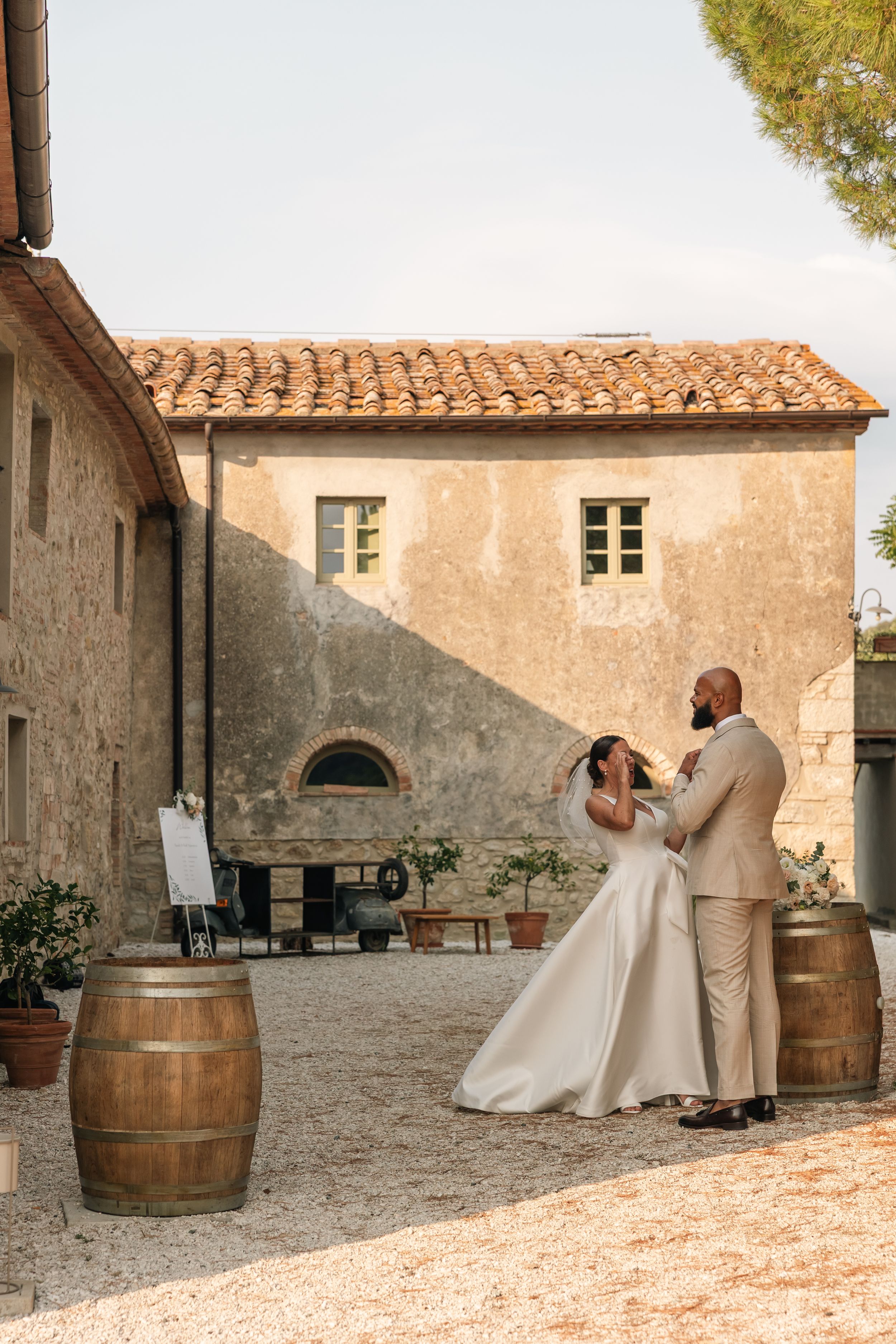 A couple shares an intimate moment in front of a rustic Italian villa with terracotta roof and wooden wine barrels.