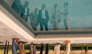 A group of people reflected in a mirrored ceiling above ornate architectural columns.