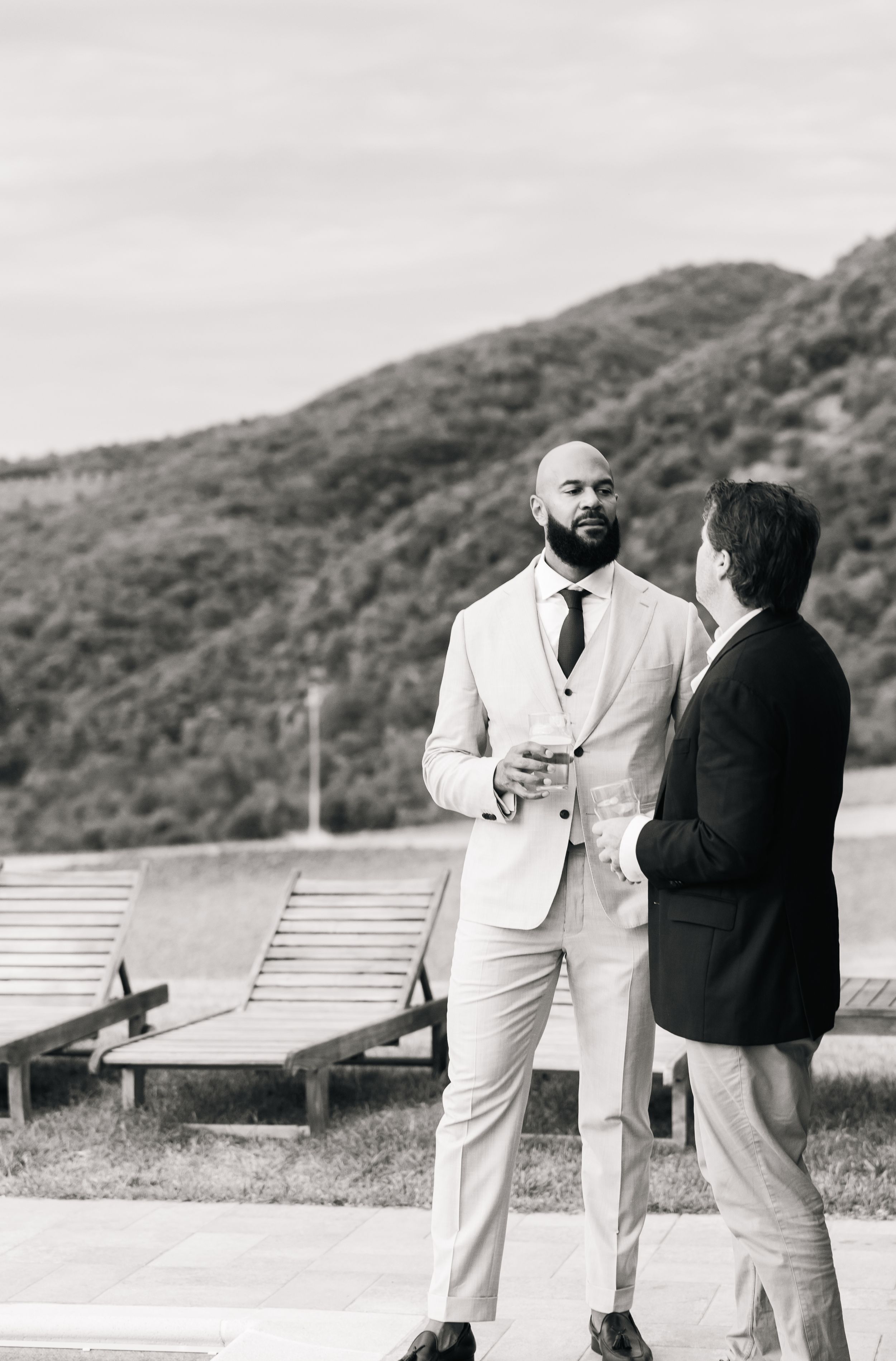 Two people in formal attire having a conversation while standing by lounge chairs with mountains in the background.