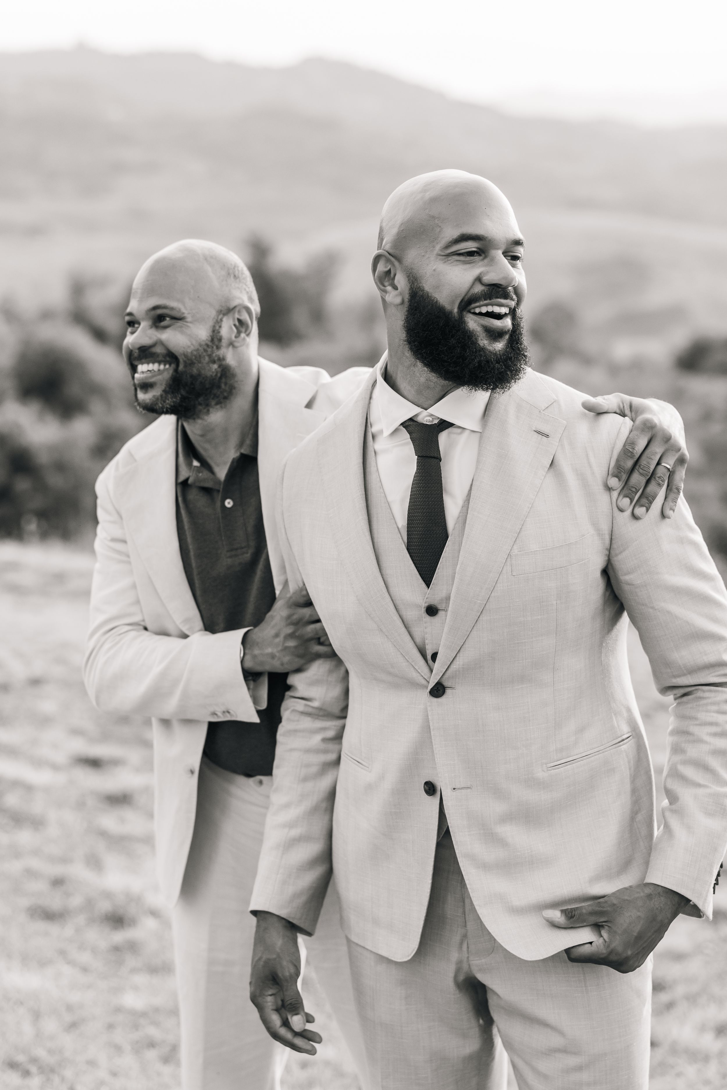 Black and white photo sequence showing two groomsmen in light suits laughing and embracing outdoors.