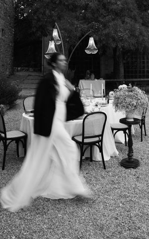 A blurred motion figure in a white dress moves past elegant outdoor table settings with hanging lanterns at dusk.