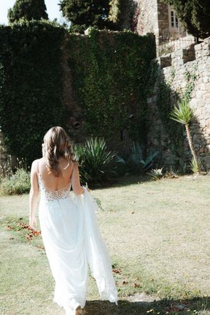 A figure in a flowing white dress walks through a sunlit garden with stone walls and greenery.