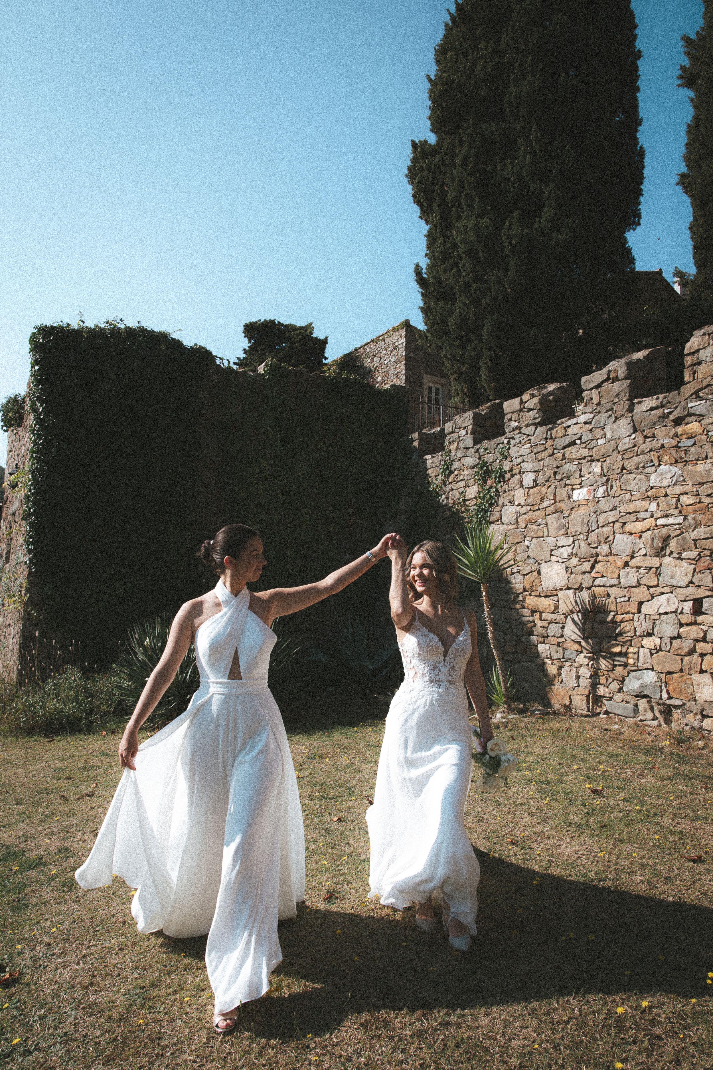 Two figures in flowing white dresses dance and twirl together against an ancient stone wall backdrop in a garden setting.
