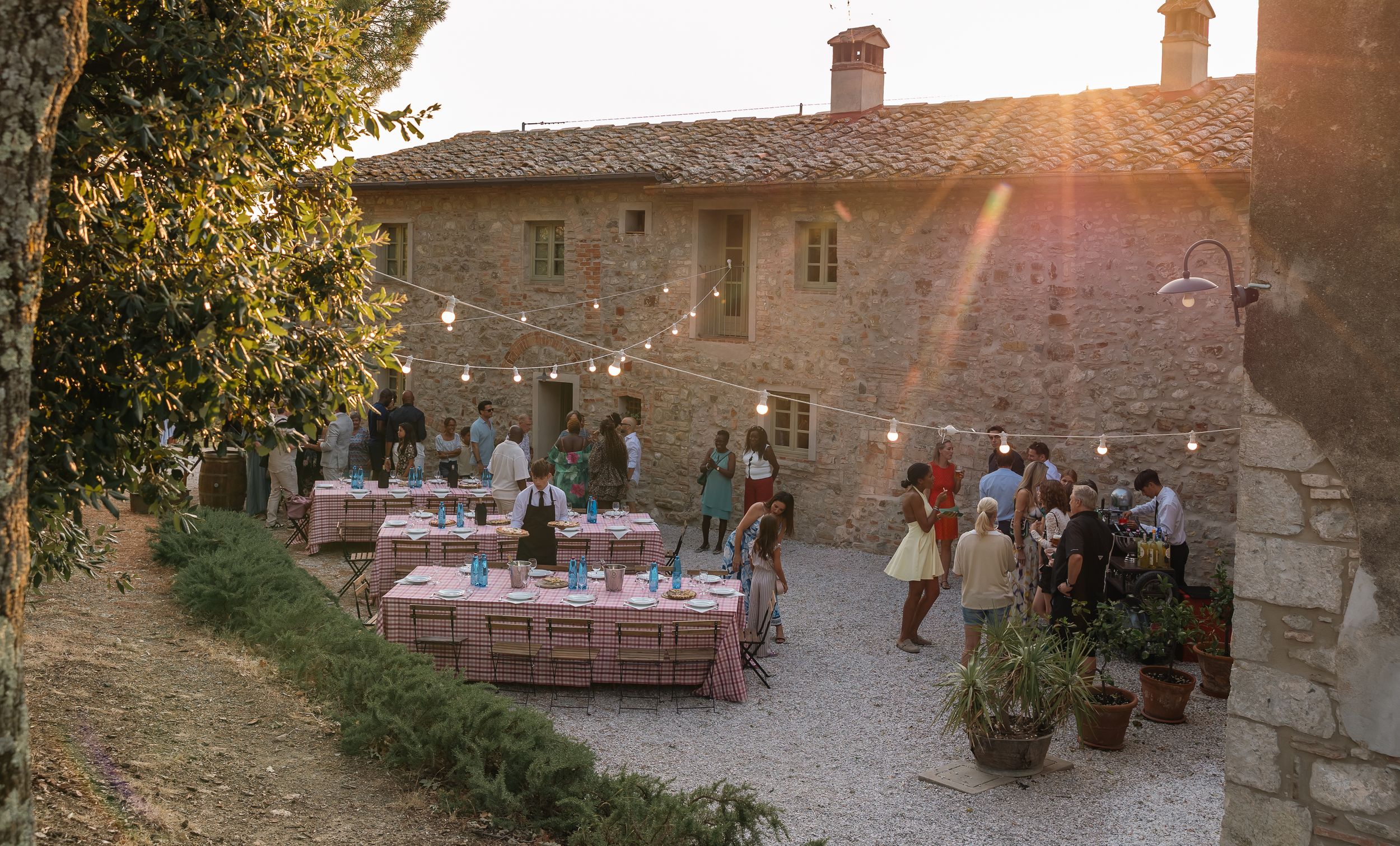 A rustic outdoor dining setup at a Tuscan villa with long wooden tables and string lights at sunset.