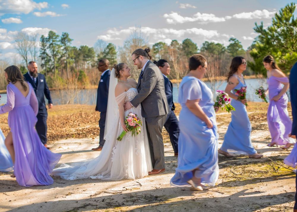 Bridal party wearing purple and blue dresses poses by lakeside at outdoor wedding ceremony on sunny day.