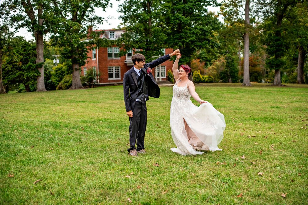 A couple in wedding attire dances playfully in a grassy field on a sunny day.