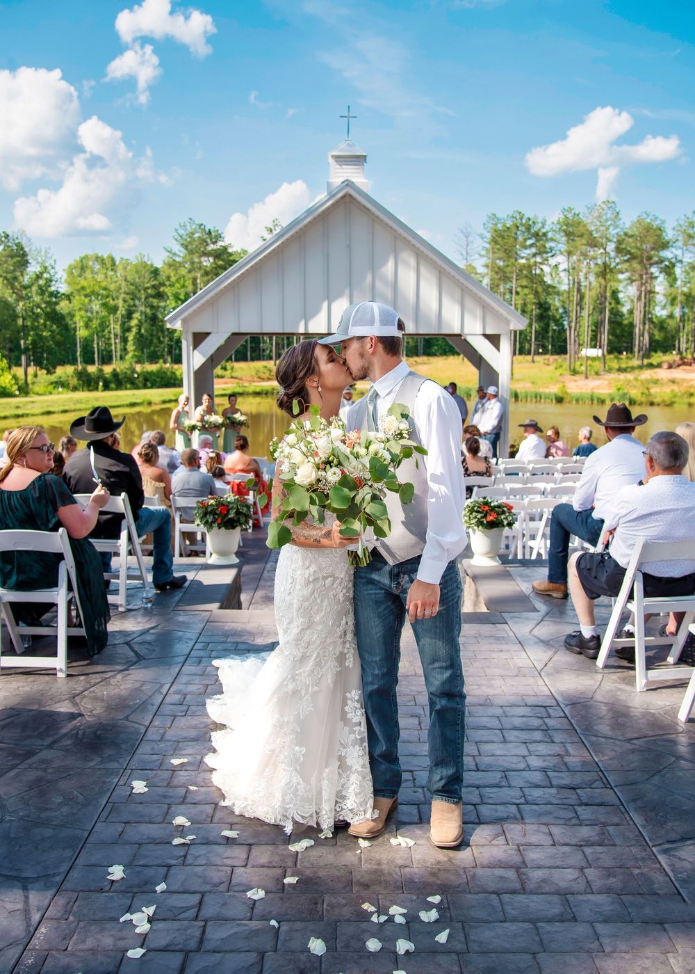 A couple shares a romantic moment under a white wedding chapel gazebo while guests look on during an outdoor ceremony.