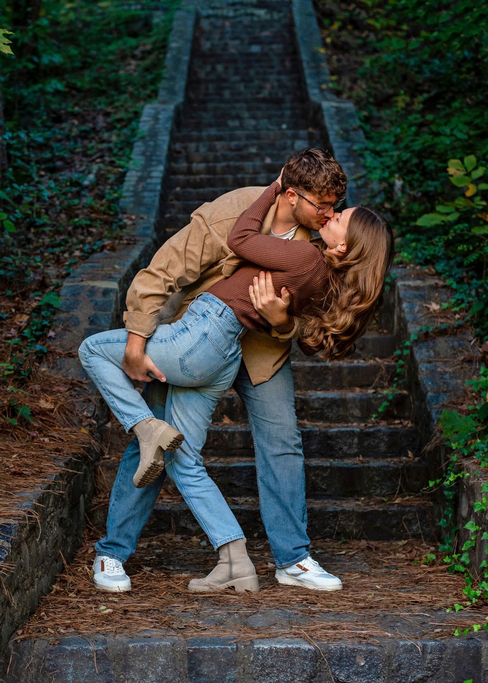 A romantic couple shares a passionate dip kiss on outdoor stone steps surrounded by greenery.
