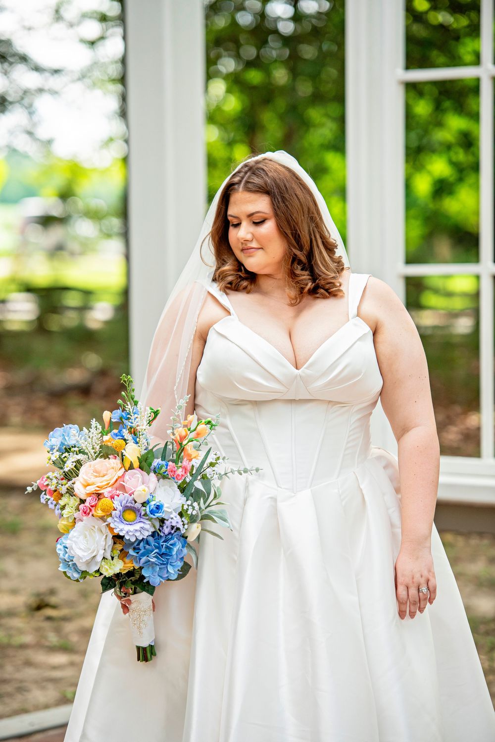 Bride in white ball gown with veil holds colorful bouquet of blue, pink, and yellow flowers outdoors.