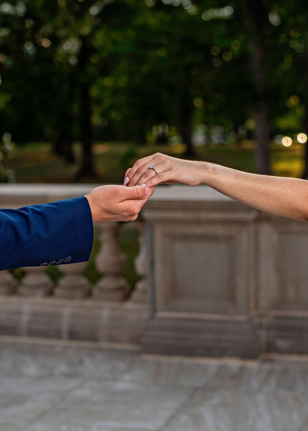 Two hands gently touching, one wearing an engagement ring, with a park and stone balustrade in the background.