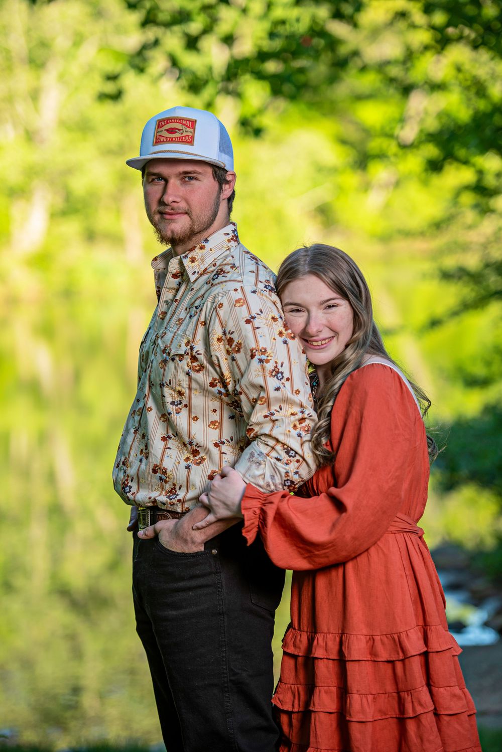 A young couple poses outdoors, woman in red dress hugging man in floral shirt and trucker hat, green trees behind.