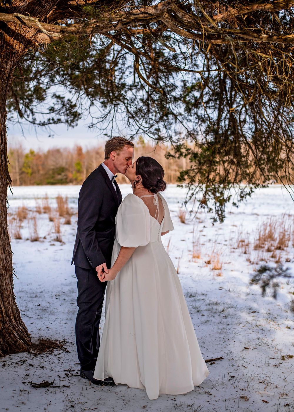 A romantic winter wedding couple shares a tender kiss under a snow-covered evergreen tree in a snowy landscape.