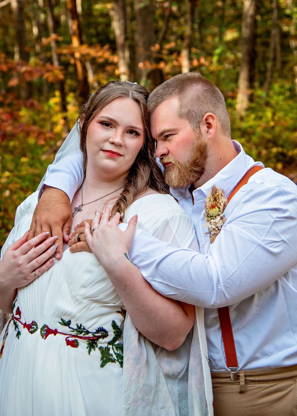 A romantic couple embraces in an autumn forest setting wearing white and rustic attire with red floral accents.