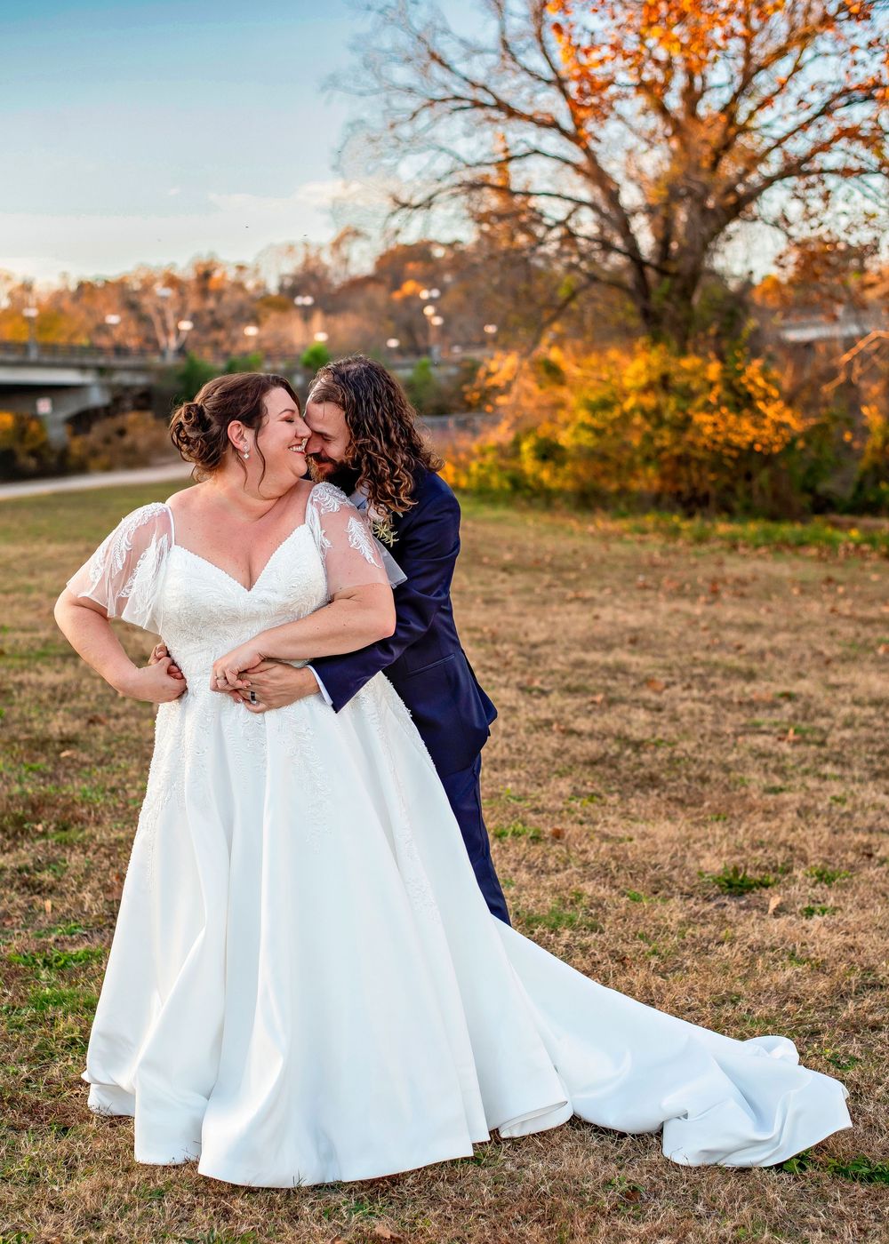 Two people in formal attire share a romantic moment in an autumn field with golden trees in the background.