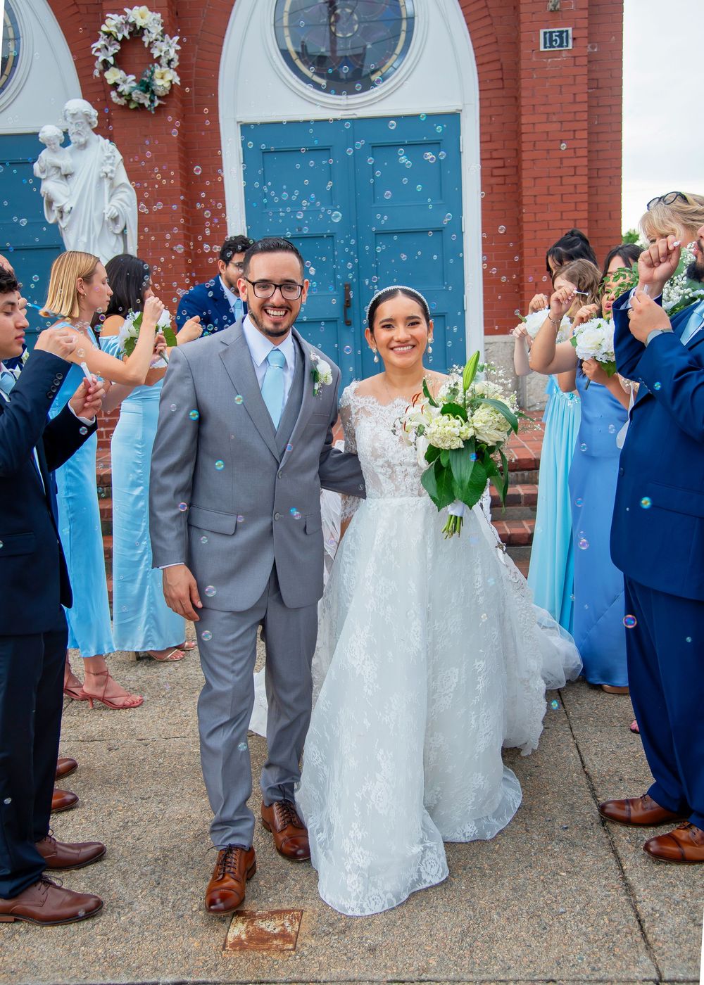 Wedding party members in light blue and gray attire stand outside a brick building with bright blue doors.