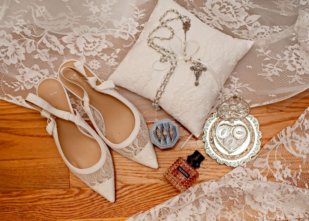 Lace wedding dress details with pearl embellishments and decorative beading lay beside elegant ivory heels on wooden floor.