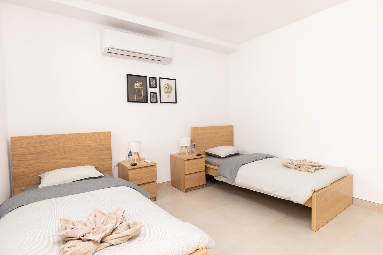 Modern twin bedroom with wooden beds, shared nightstand, air conditioning unit, and white walls.