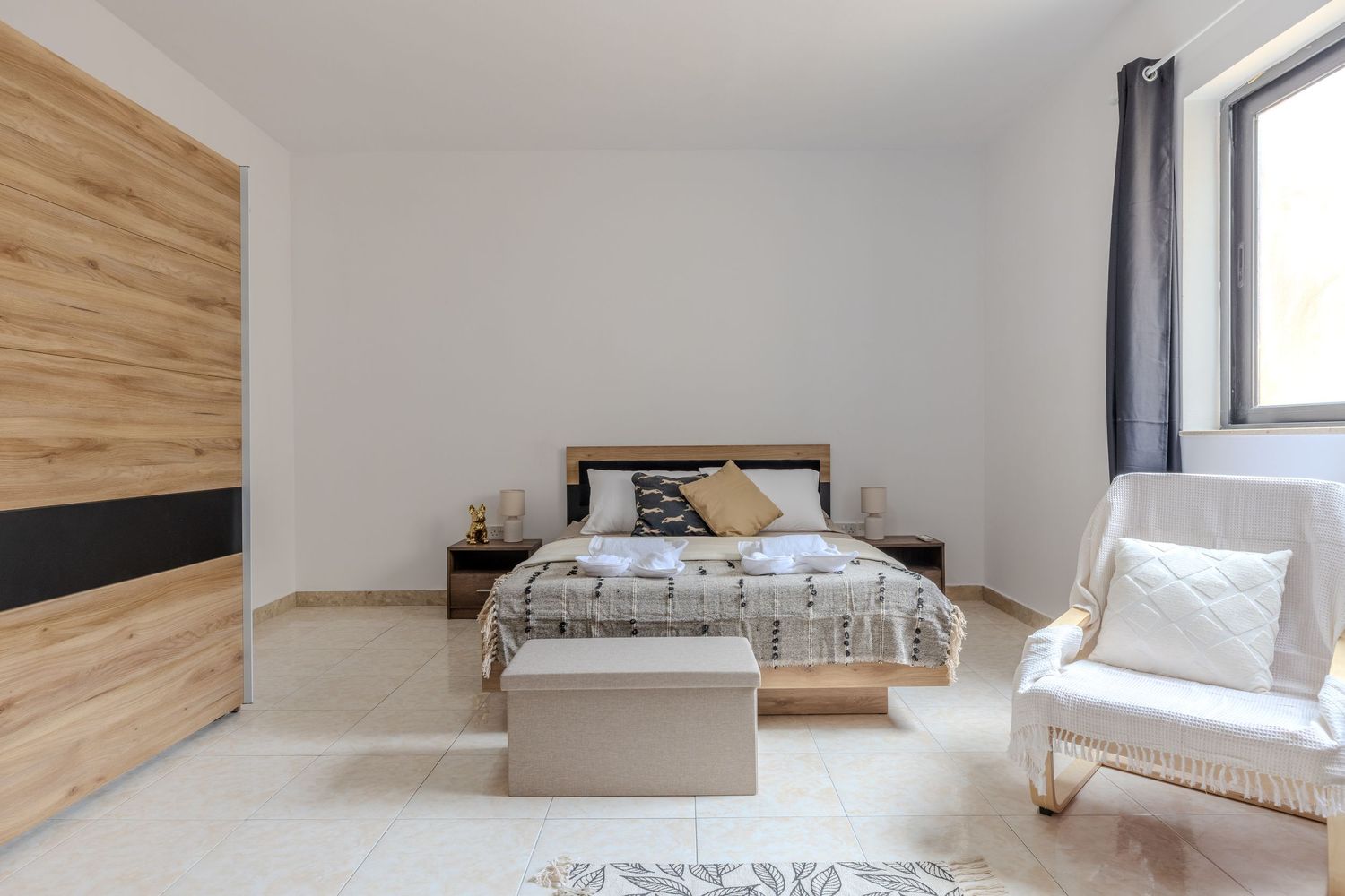 Bright minimalist bedroom with wooden wardrobe, double bed, white armchair, and natural window light.