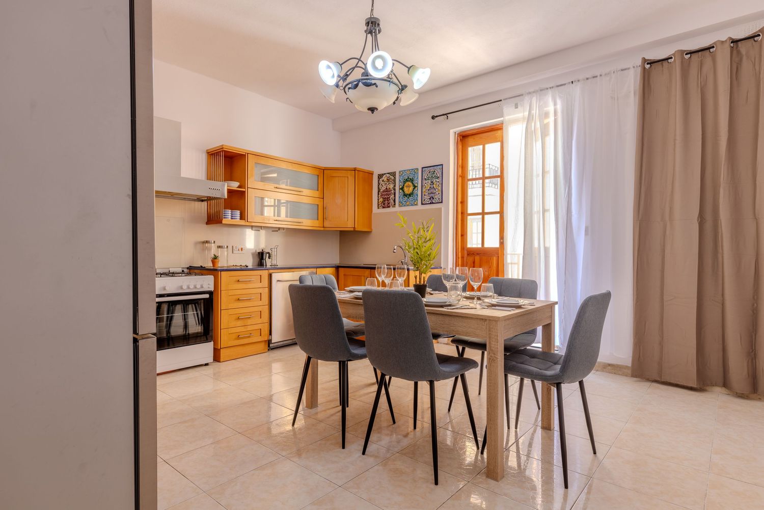 Charming kitchen-dining area with wooden cabinets, chandelier, gray chairs, and natural window light.