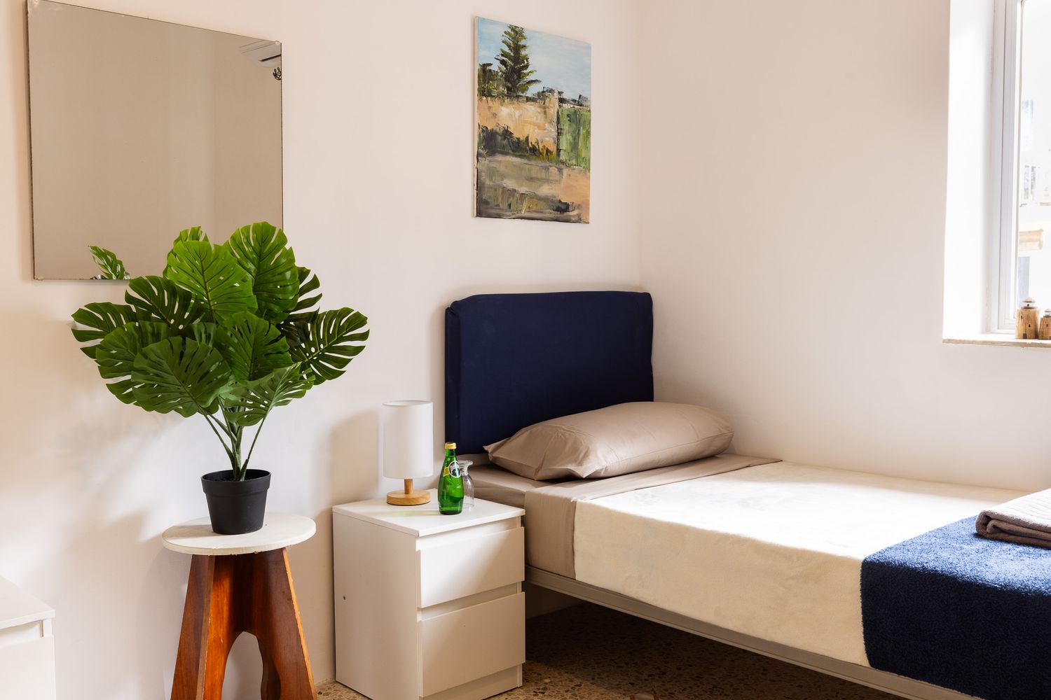 Minimalist single bedroom with navy headboard, monstera plant, mirror, and landscape painting on white wall.