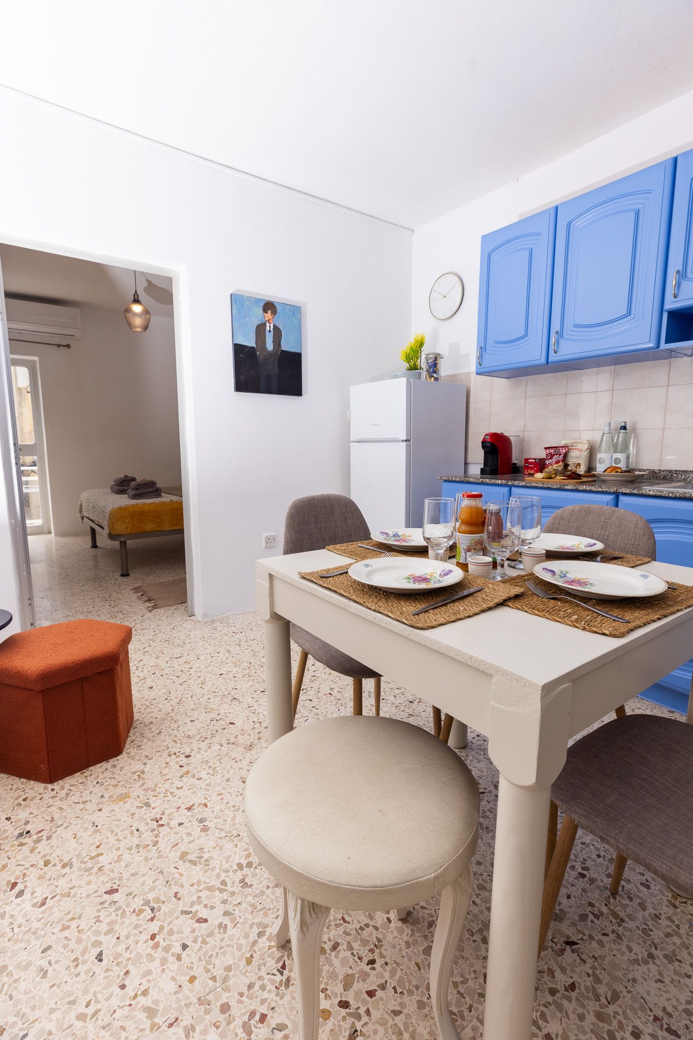 Open-plan apartment with blue kitchen cabinets, white dining table, terrazzo floors, and bedroom view.