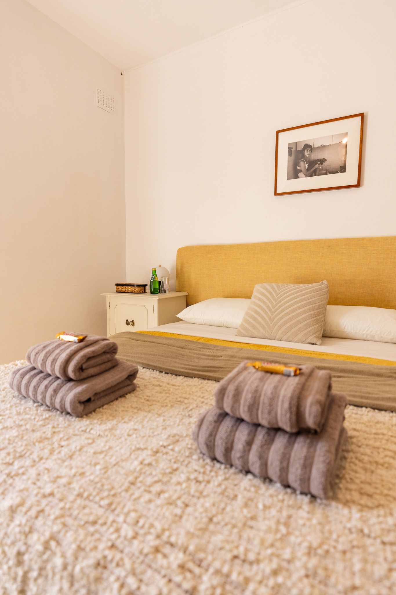 Warm bedroom with yellow upholstered bed, striped folded towels, shag rug, and framed black-white photo.