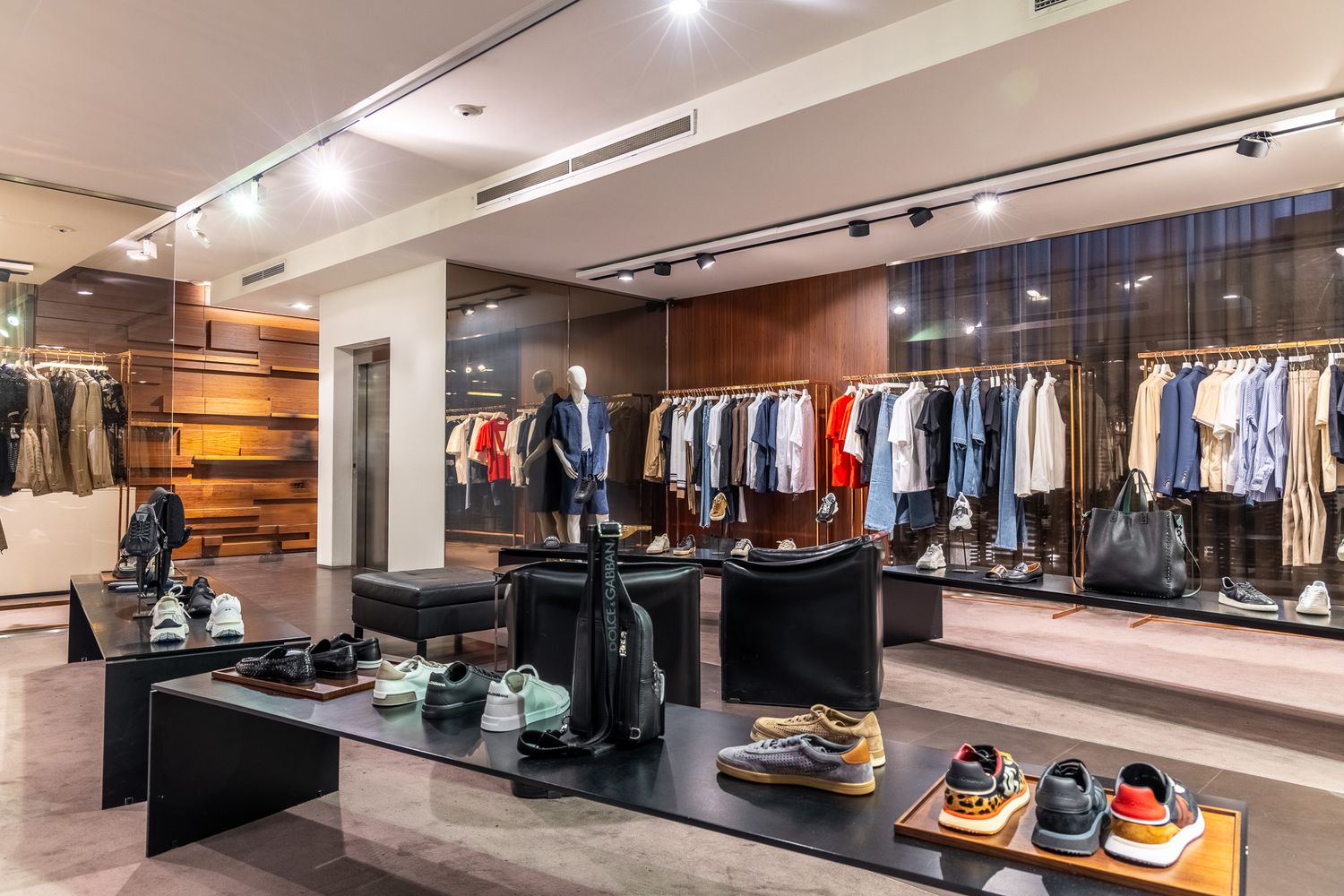 Modern menswear store with sneakers on display tables, clothing racks, and wood-paneled walls.